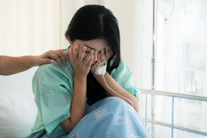 一名妇女坐在医院病床上，用手捂住脸，她伴侣的手搭在她的肩膀上。